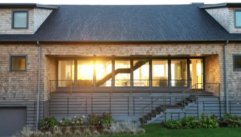 Cedar shingle siding on coastal New England home exterior with large glass windows and sunset light reflecting through interior